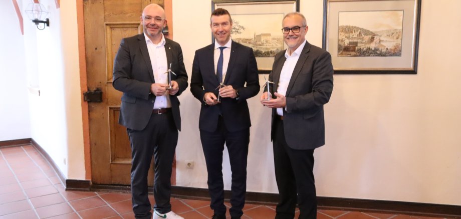 3 Männer in einer historischen Räumlichkeit; sie halten jeder eine Modell-Windkraftanlage in den Händen