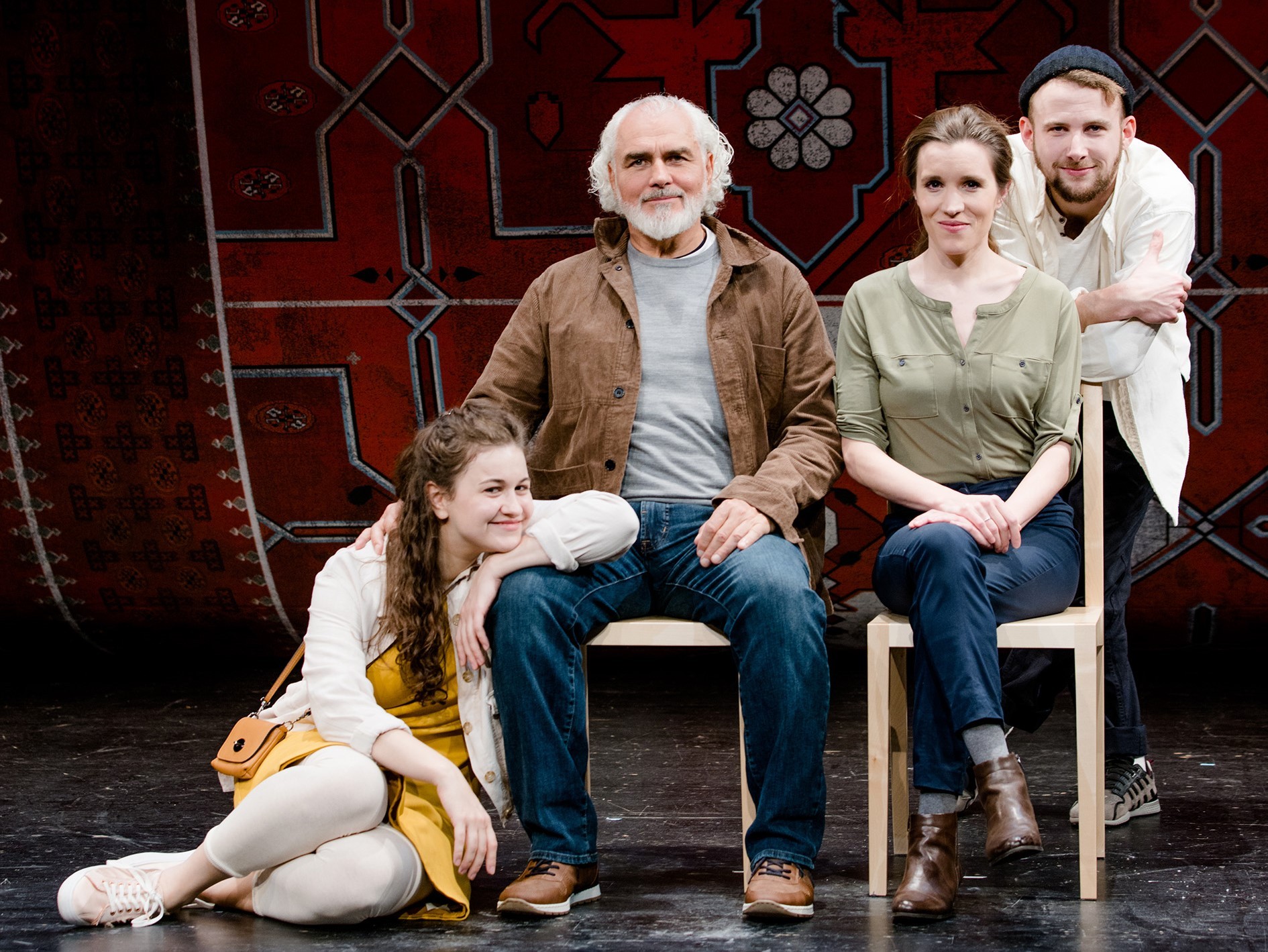 Ein Mann und eine Frau sitzen auf Stühlen, neben dem Mann sitzt eine junge Frau auf dem Boden und hinter der Frau steht ein junger Mann 